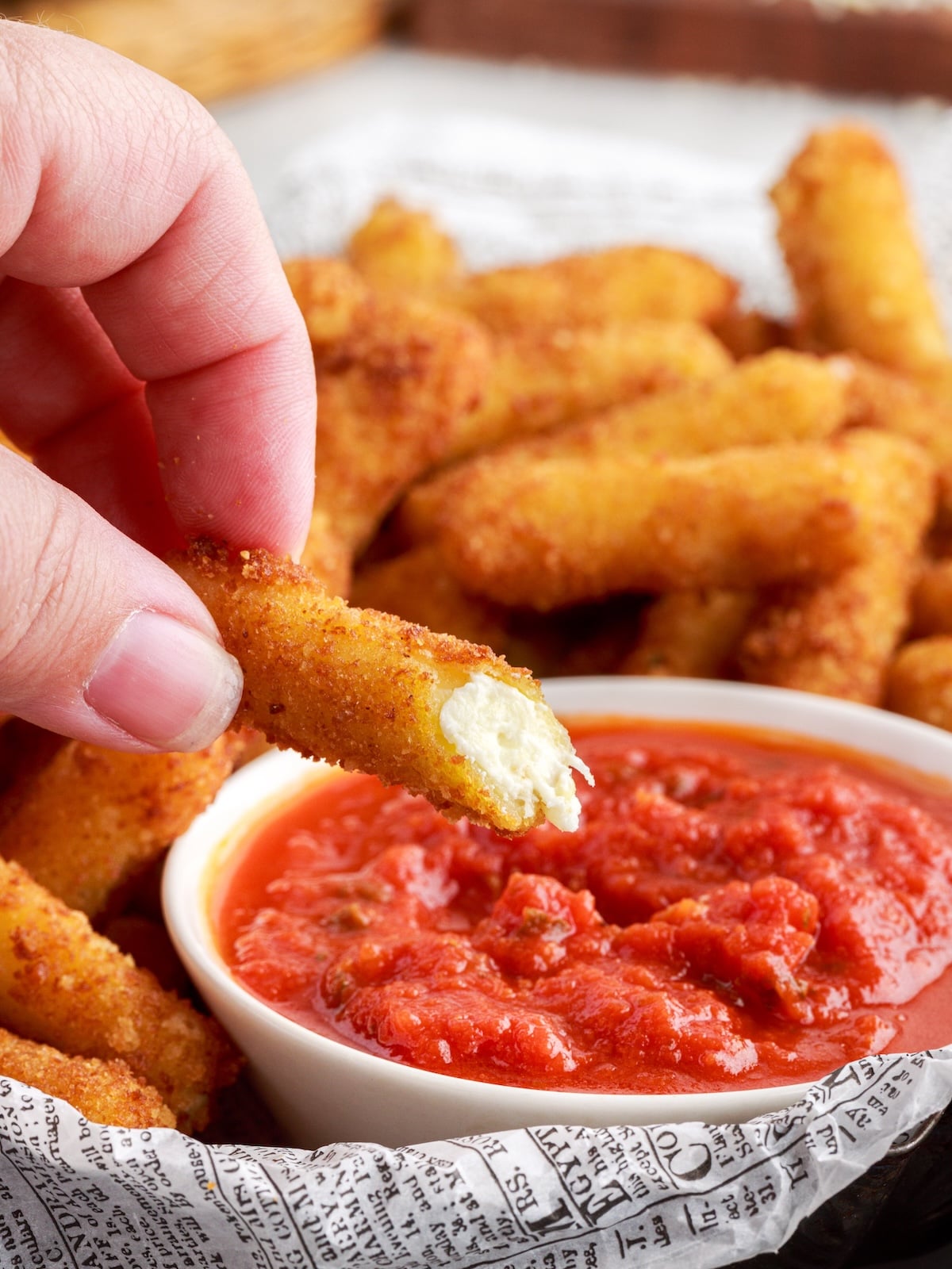 A hand holds a breaded stuffed ziti fritta dipped in marinara sauce, with more Stuffed Ziti Fritta on a tray lined with newspaper-style paper in the background.