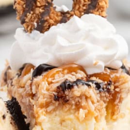Close-up of a decadent Samoa Cheesecake slice topped with whipped cream, toasted coconut, caramel, and a classic Samoa cookie; text reads SAMOA CHEESECAKE and VINTAGE RECIPE!.