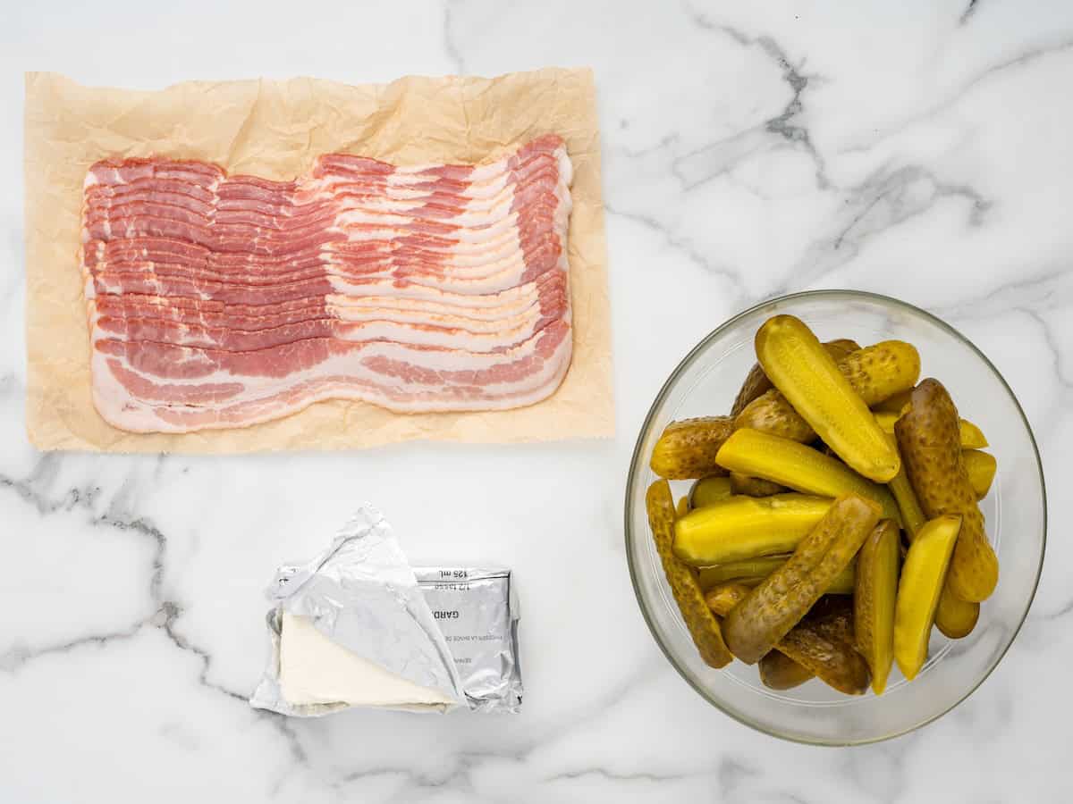 Raw bacon on parchment, a glass bowl filled with whole pickles, and a block of unwrapped cream cheese sit on a white marble surface—everything you need to make delicious Bacon Wrapped Pickles.