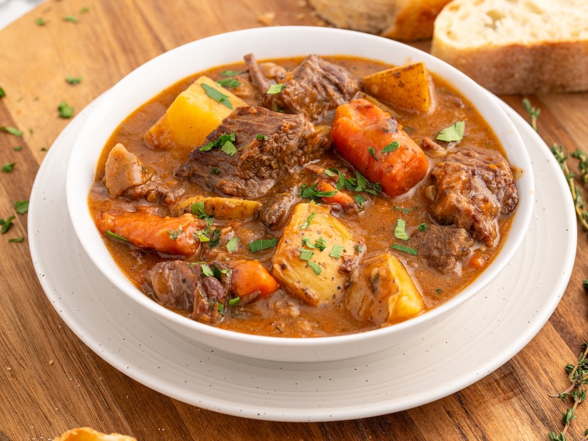 A bowl of hearty Beef Brisket Stew with chunks of beef, potatoes, carrots, and herbs in a rich broth, served on a white plate with slices of bread on a wooden surface.