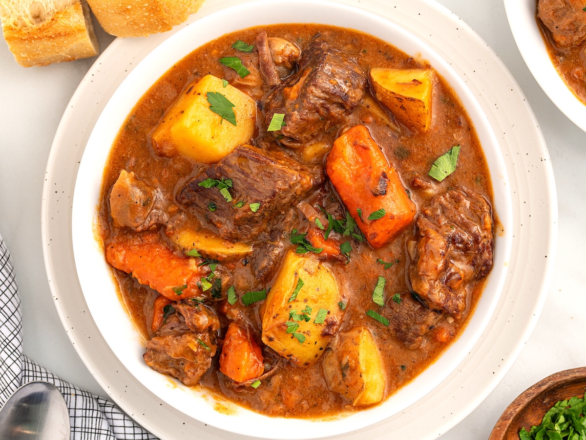 A bowl of Beef Brisket Stew with large chunks of beef, potatoes, and carrots in a rich brown broth, garnished with chopped parsley. Slices of bread are visible in the background.