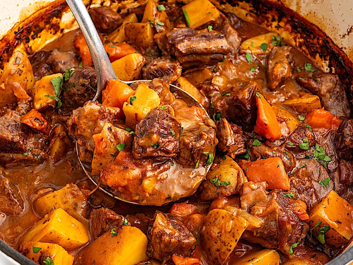 A close-up of a hearty Beef Brisket Stew with tender chunks of beef, potatoes, carrots, and herbs in a rich brown sauce, being scooped with a large metal ladle from a pot.
