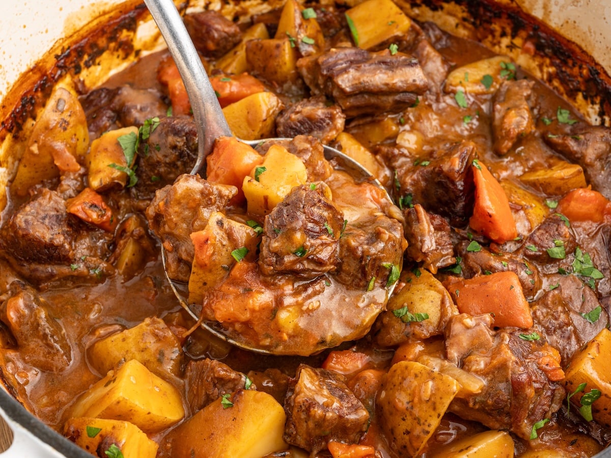 A ladle lifting tender beef brisket, golden potatoes, and carrots from a Dutch oven filled with rich, savory beef brisket stew.