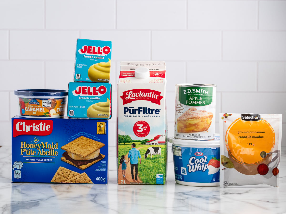 A variety of food items on a kitchen counter, perfect for making a Caramel Apple Eclair Cakeโfeaturing Jello pudding, Lactantia milk, E.D. Smith apple pie filling, Cool Whip, Honey Maid graham crackers, Selection caramel sauce, and Philadelphia cream cheese.
