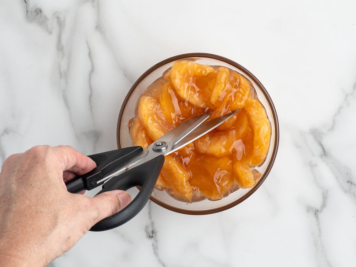 A hand uses kitchen scissors to cut canned peach slices in syrup inside a glass bowl, set on a white marble surfaceโperfect for topping a Caramel Apple Eclair Cake or other creative desserts.