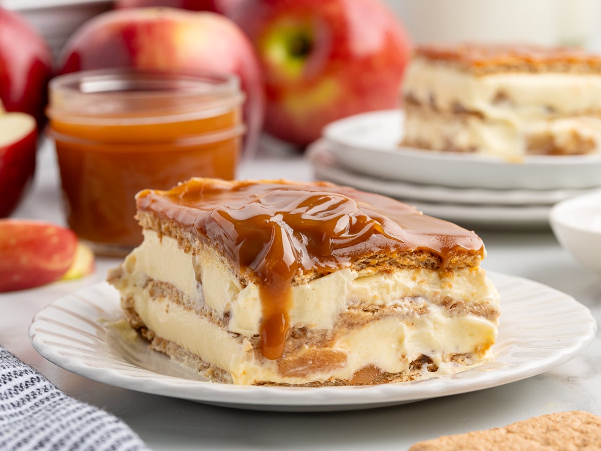 A slice of creamy Caramel Apple Eclair Cake topped with caramel sauce sits on a white plate, with apples, caramel in a jar, and another dessert slice blurred in the background.