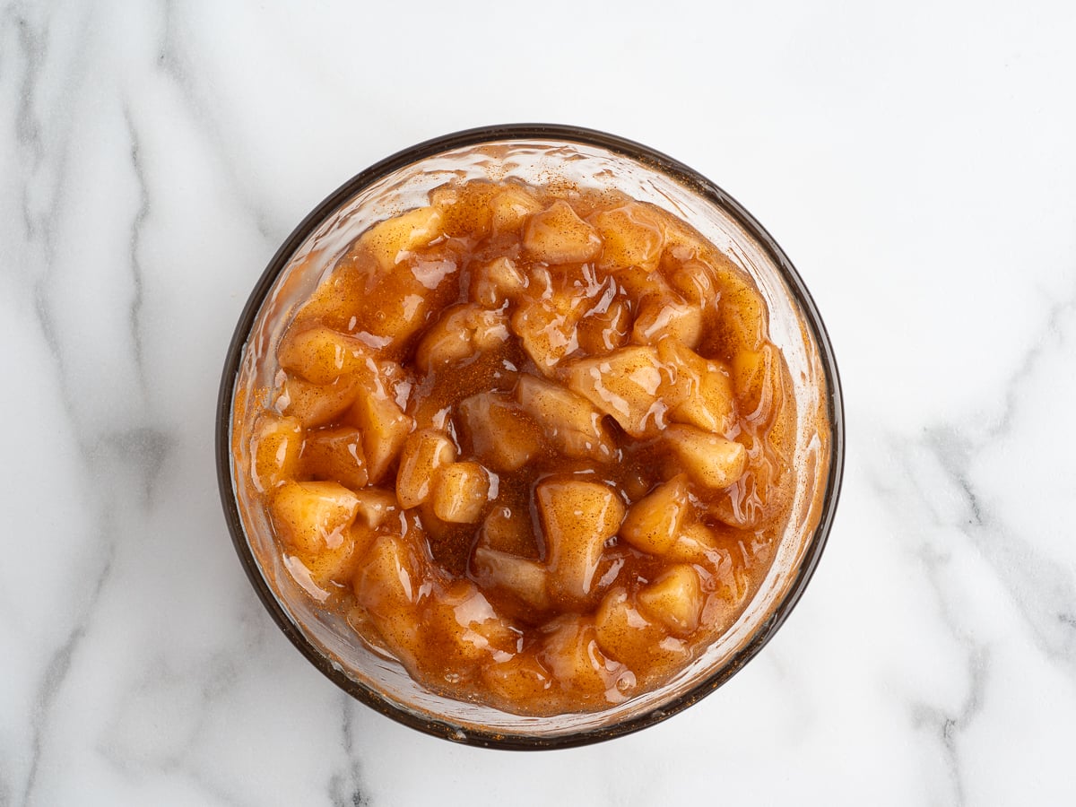 A glass bowl filled with chunky apple pie filling, coated in a glossy brown cinnamon sauce, sits on a white marble surfaceโperfect for topping a Caramel Apple Eclair Cake.
