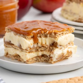A delicious Eclair Cake made of layers of graham crackers and creamy filling, topped with caramel sauce, is served on a white plate. A jar of caramel and fruit are blurred in the background.