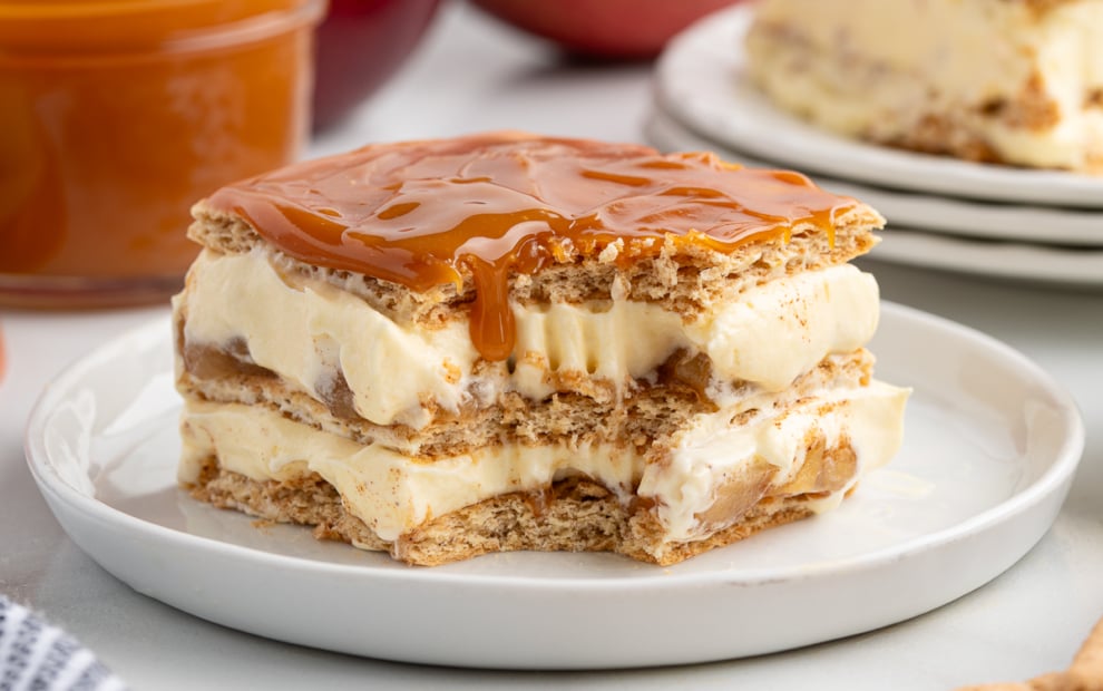 A delicious Eclair Cake made of layers of graham crackers and creamy filling, topped with caramel sauce, is served on a white plate. A jar of caramel and fruit are blurred in the background.