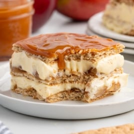 A square slice of Caramel Apple Eclair Cake with creamy vanilla filling and caramel sauce on top, served on a white plate. The layers appear to be crispy wafers and smooth cream.
