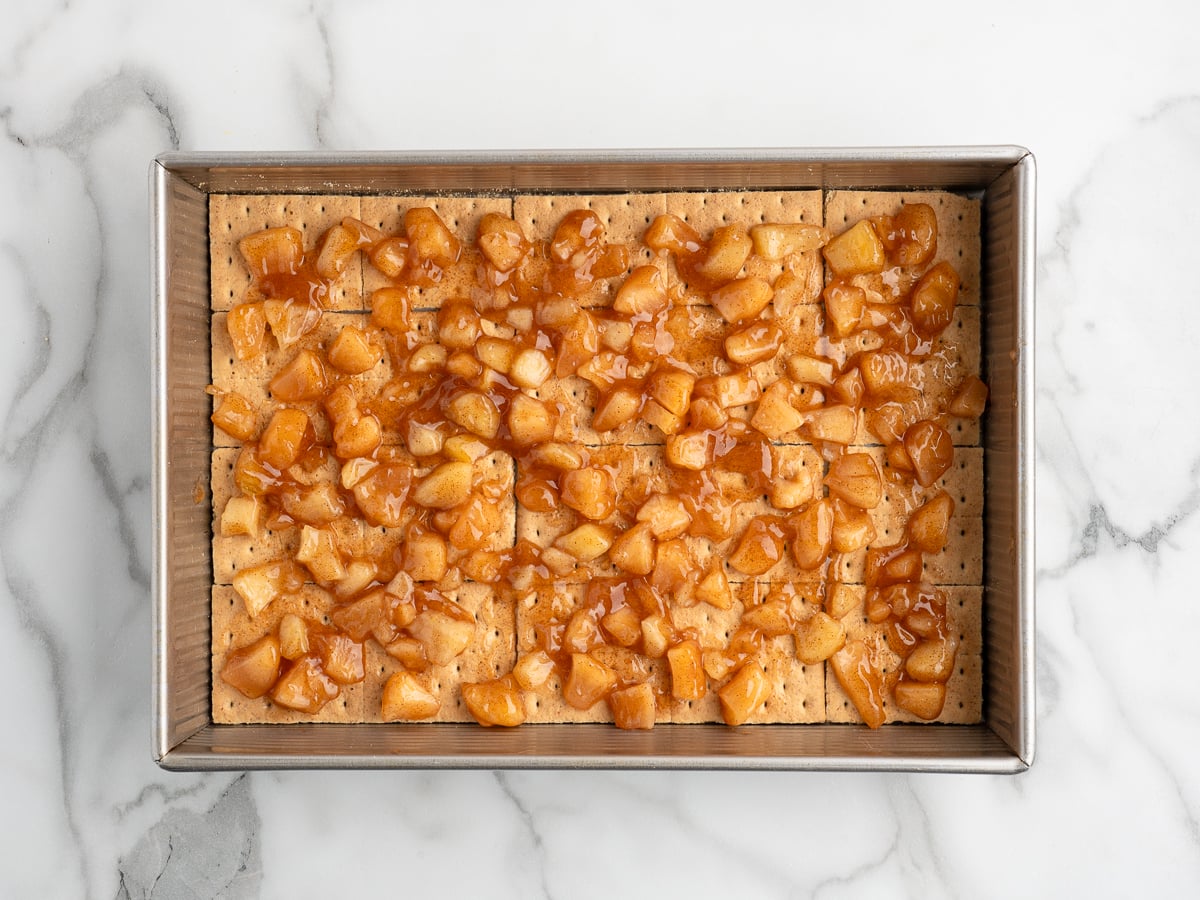 A rectangular baking pan filled with a layer of graham crackers, topped with diced apples coated in a glossy cinnamon-sugar mixture, brings this Caramel Apple Eclair Cake to life on a white marble surface.