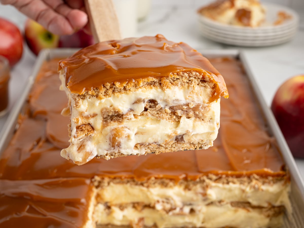 A close-up of a creamy layered Eclair Cake with caramel topping being lifted from a baking pan. The dessert has visible layers of cream, cookies or cake, and caramel. Apples and plates are visible in the background.