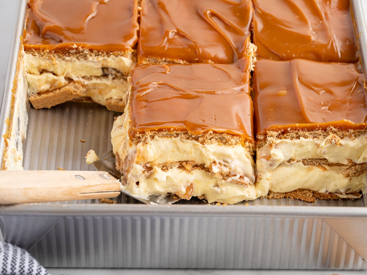 A close-up of a rectangular pan with several slices of Caramel Apple Eclair Cake featuring a caramel topping, creamy filling, and graham cracker layers. One piece is being lifted with a metal spatula.