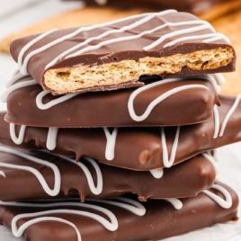 A stack of chocolate-covered graham crackers with white chocolate drizzle, set against a background of whole graham crackers.