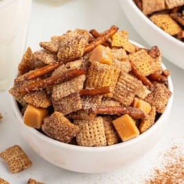 Bowl of churro Chex Mix with pretzels and caramel squares, coated in cinnamon sugar, next to a glass of milk.