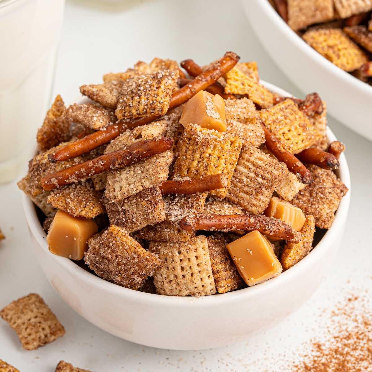 Bowl of churro Chex Mix with pretzels and caramel squares, coated in cinnamon sugar, next to a glass of milk.