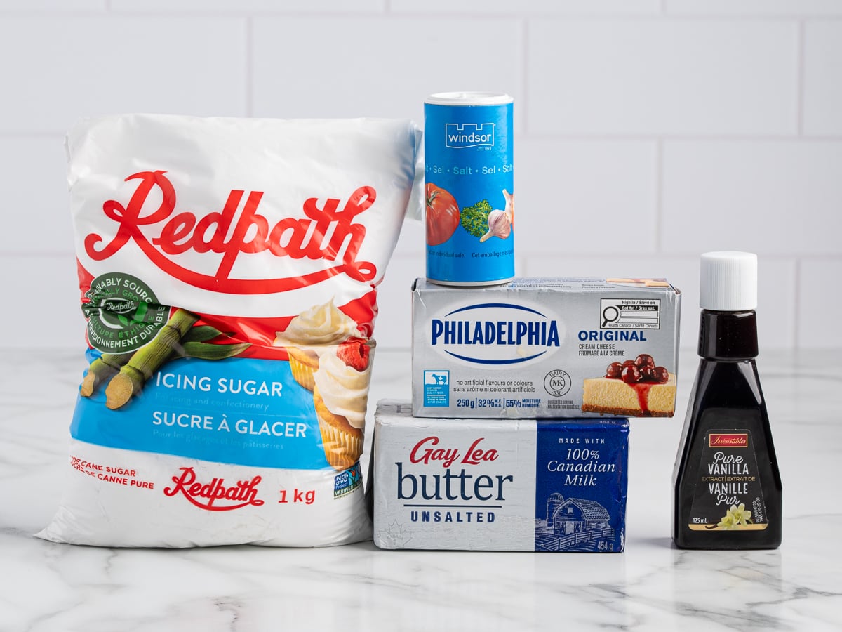 A group of baking ingredients on a marble counter, perfect for Cream Cheese Frosting: a 1 kg bag of Redpath icing sugar, Windsor salt shaker, Philadelphia cream cheese, Gay Lea unsalted butter, and a bottle of vanilla extract.