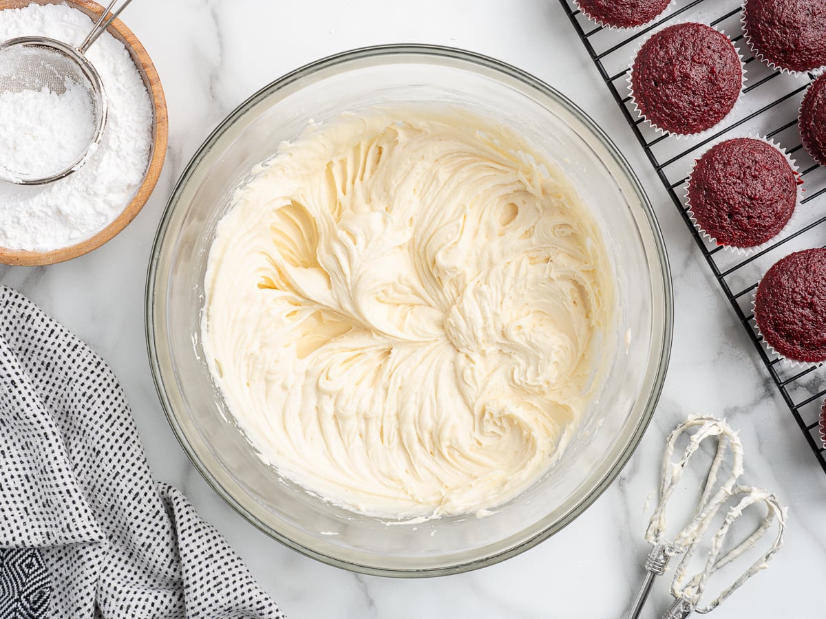 A glass bowl filled with luscious Cream Cheese Frosting sits on a marble counter, surrounded by powdered sugar, a patterned towel, a whisk, and freshly baked red velvet cupcakes cooling on a wire rack.
