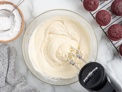 A bowl of creamy cream cheese frosting is whipped with a hand mixer, surrounded by powdered sugar, a sifter, a kitchen towel, and red velvet cupcakes cooling on a wire rack.