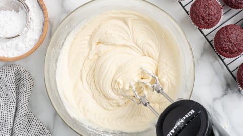 A bowl of creamy cream cheese frosting is whipped with a hand mixer, surrounded by powdered sugar, a sifter, a kitchen towel, and red velvet cupcakes cooling on a wire rack.