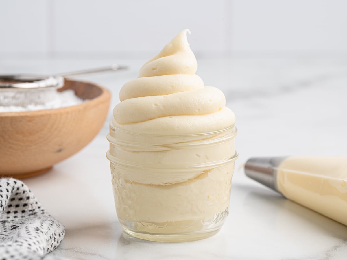 A small glass jar filled with swirled cream cheese frosting sits on a marble surface, with a piping bag, a bowl of powdered sugar, and a cloth nearby.