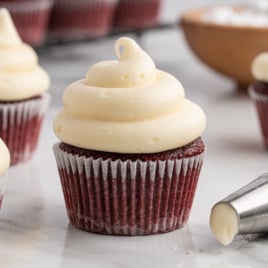 A red velvet cupcake in a white paper liner is topped with a tall swirl of cream cheese frosting. Other frosted cupcakes, a piping bag filled with cream cheese frosting, and a bowl of powdered sugar are visible in the background.