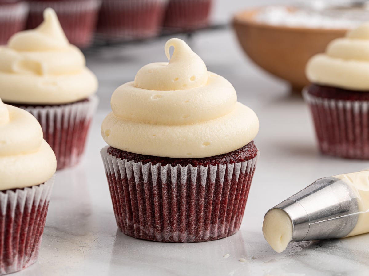 A red velvet cupcake in a white paper liner is topped with a tall swirl of cream cheese frosting. Other frosted cupcakes, a piping bag filled with cream cheese frosting, and a bowl of powdered sugar are visible in the background.