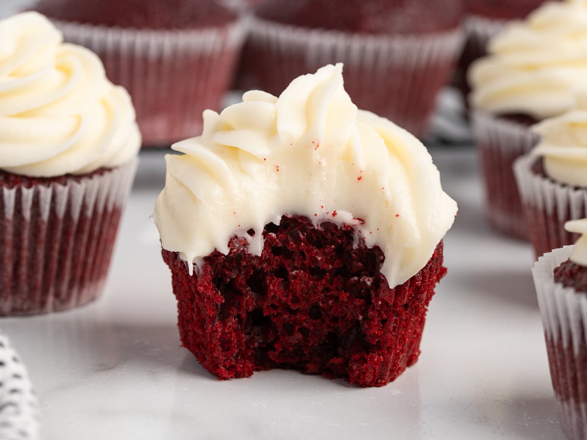 A red velvet cupcake with rich cream cheese frosting sits on a white surface. A large bite reveals its moist red interior, while other cupcakes topped with cream cheese frosting are blurred in the background.