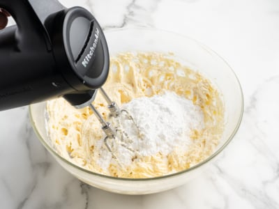 A hand mixer is blending powdered sugar into a creamy cream cheese frosting mixture in a glass bowl on a white marble surface.
