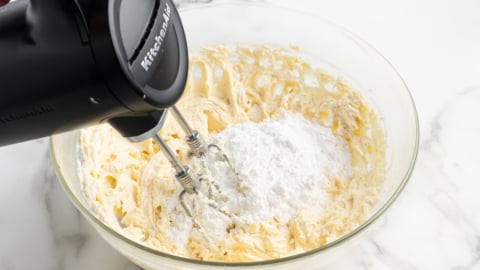 A hand mixer is blending powdered sugar into a creamy cream cheese frosting mixture in a glass bowl on a white marble surface.