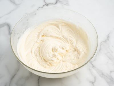 A glass bowl filled with smooth, creamy white cream cheese frosting sits on a marble countertop. The frosting has elegant swirled patterns from mixing.