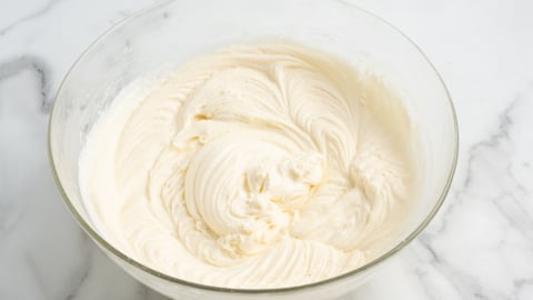 A glass bowl filled with smooth, creamy white cream cheese frosting sits on a marble countertop. The frosting has elegant swirled patterns from mixing.