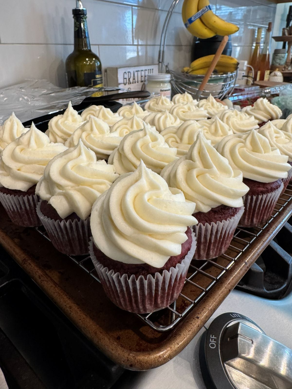 A tray of red velvet cupcakes with swirled cream cheese frosting sits on a wire rack in a kitchen, surrounded by various kitchen items like bananas and bottles in the background.