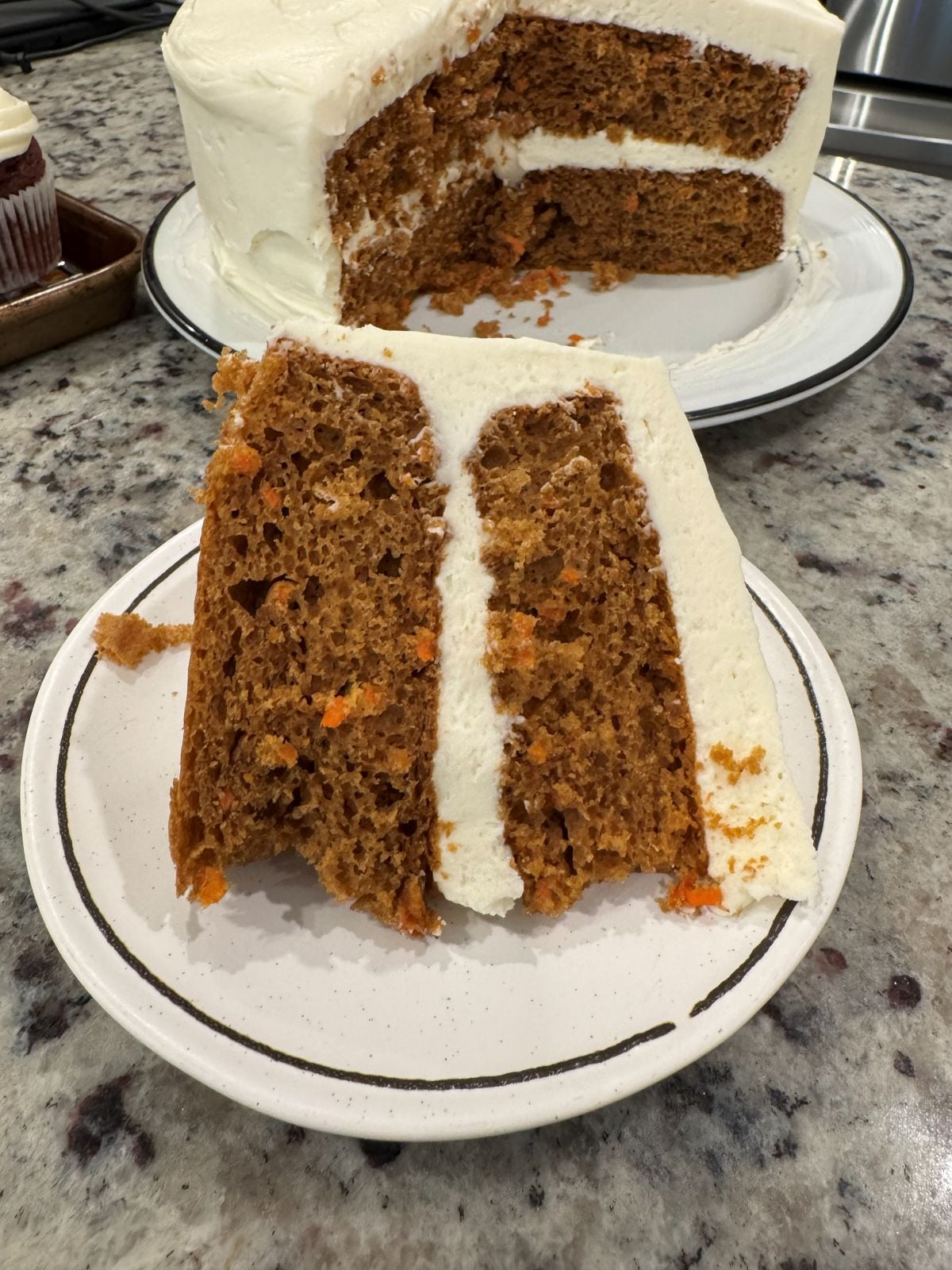 A slice of two-layer carrot cake with rich cream cheese frosting is served on a white plate. The whole cake, missing one slice, sits in the background on a white platter atop a granite countertop.