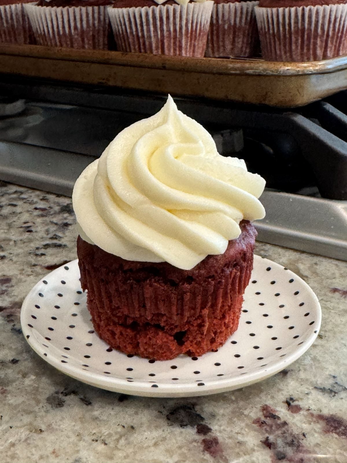 A red velvet cupcake with cream cheese frosting sits on a white plate with black polka dots, on a kitchen counter. More cupcakes are visible on a baking tray in the background.