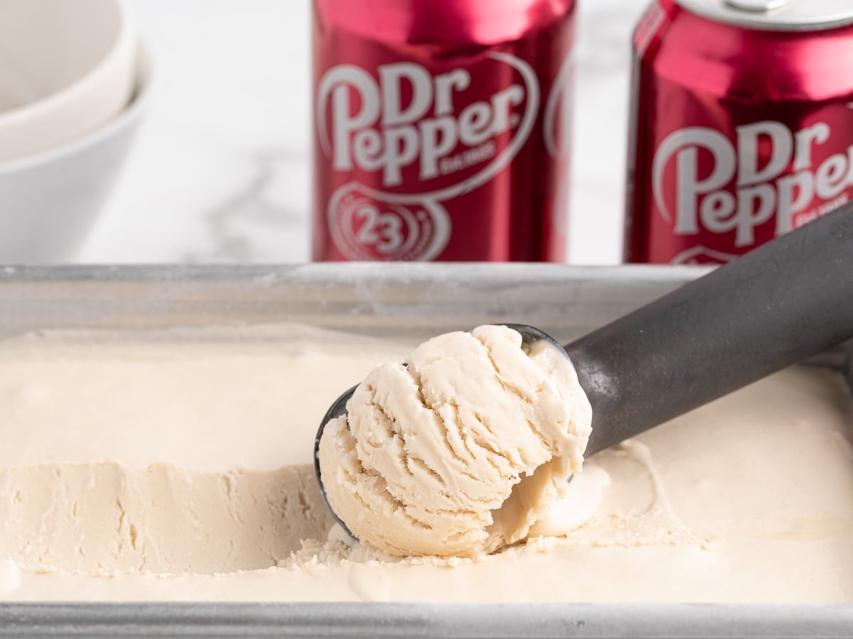 A close-up of a metal ice cream scoop lifting a scoop of creamy Dr Pepper Ice Cream from a rectangular pan, with two red Dr Pepper cans and white bowls in the background.