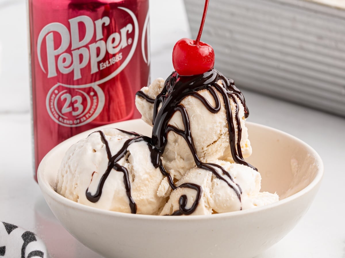 A bowl of vanilla Dr Pepper Ice Cream topped with chocolate syrup and a cherry, with a can of Dr Pepper soda in the background.