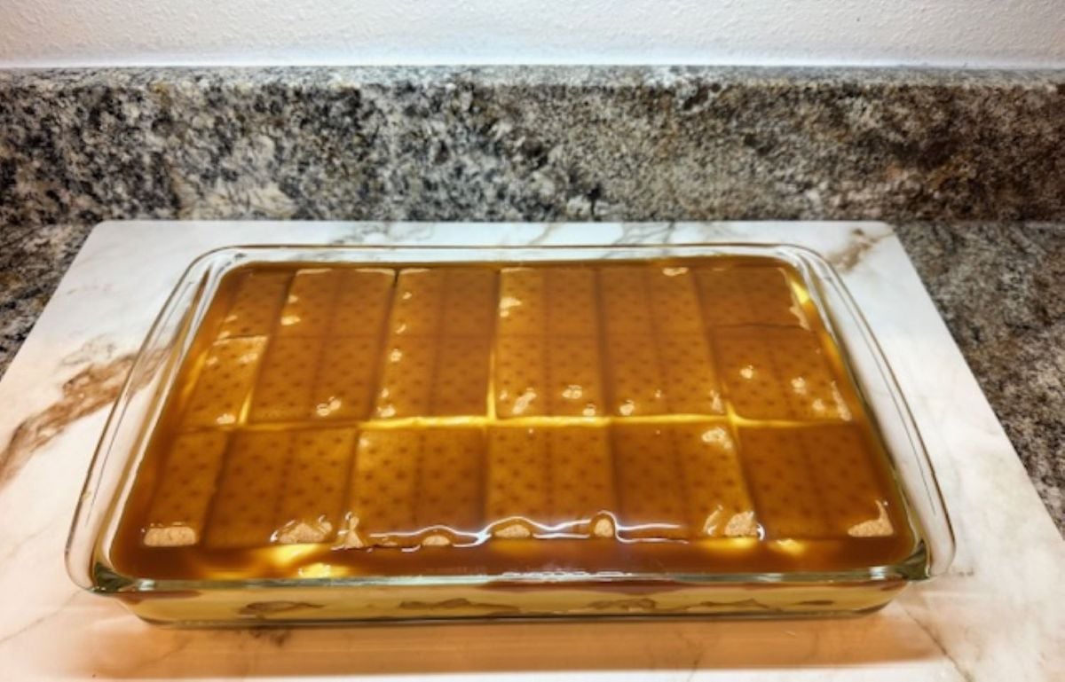 A glass baking dish filled with a layered Eclair Cake, topped with a smooth caramel glaze and rectangular cookies arranged in neat rows on the surface, set on a marble countertop.