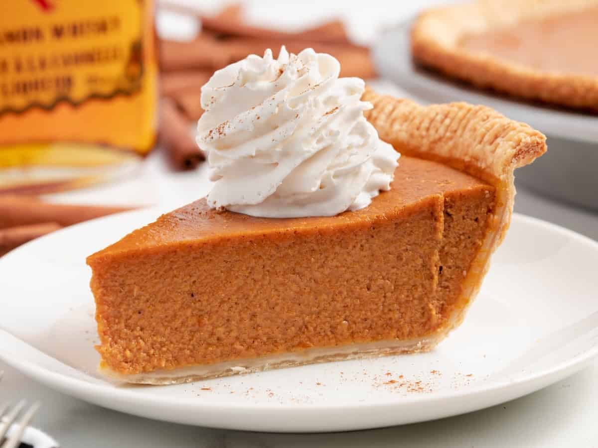 Pumpkin pie topped with a swirl of whipped cream and a dusting of cinnamon on a white plate, with a bottle of Fireball whisky, cinnamon sticks, and a whole pie blurred in the background.