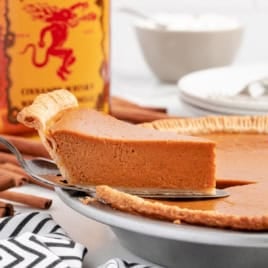 A slice of pumpkin pie being lifted from a pie pan, with cinnamon sticks and a Fireball Cinnamon Whisky bottle in the background, perfectly capturing the essence of Fireball Pumpkin Pie.