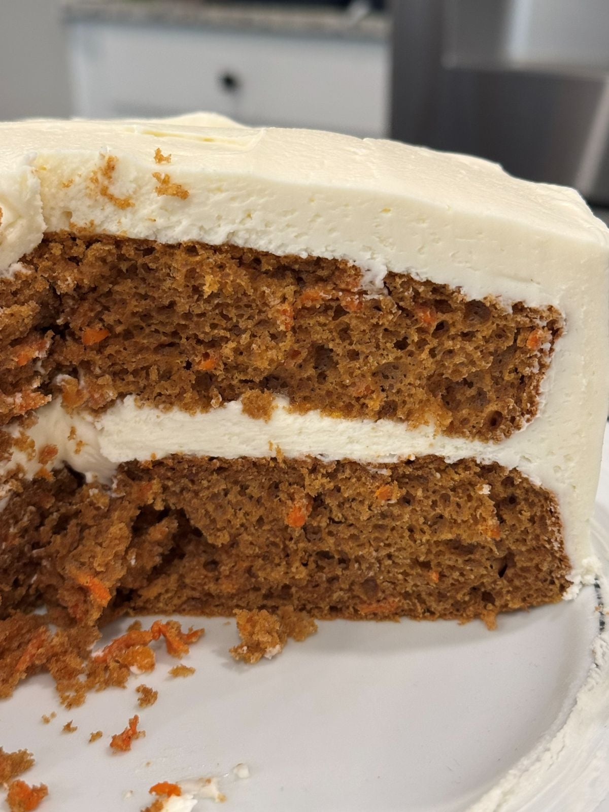 A close-up of a sliced, two-layer carrot cake with rich cream cheese frosting, showing moist cake layers with bits of carrot and a generous layer of creamy frosting between and on top.