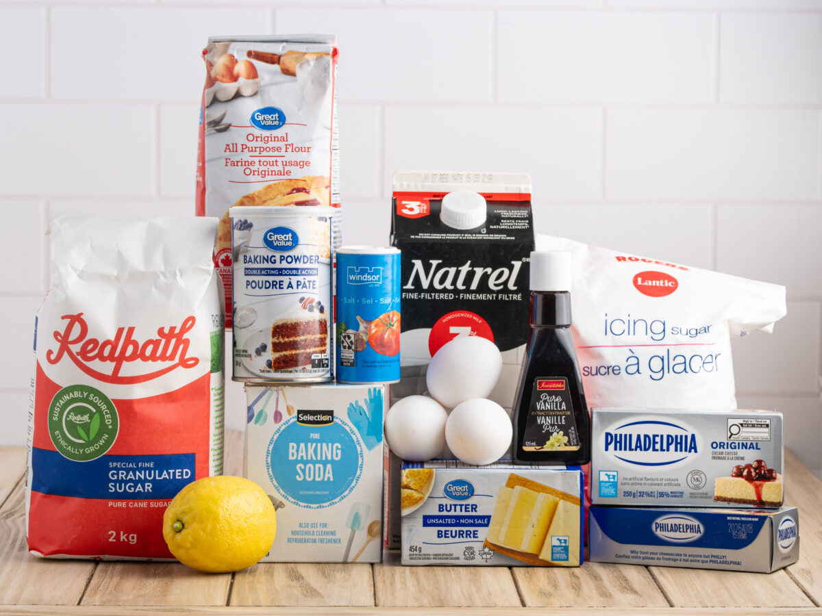 Assorted baking ingredients on a wooden surface, perfect for making Lemon Coffee Cake, including flour, sugar, icing sugar, baking powder, baking soda, butter, eggs, cream cheese, vanilla, milk, and a lemon in front of a white tile background.