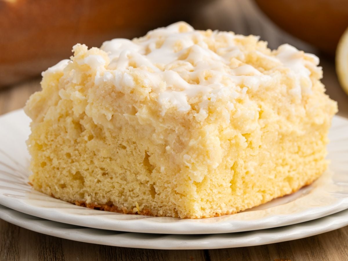 A close-up of a thick slice of crumbly, golden Lemon Coffee Cake with a light drizzle of white icing on top, served on a white plate.