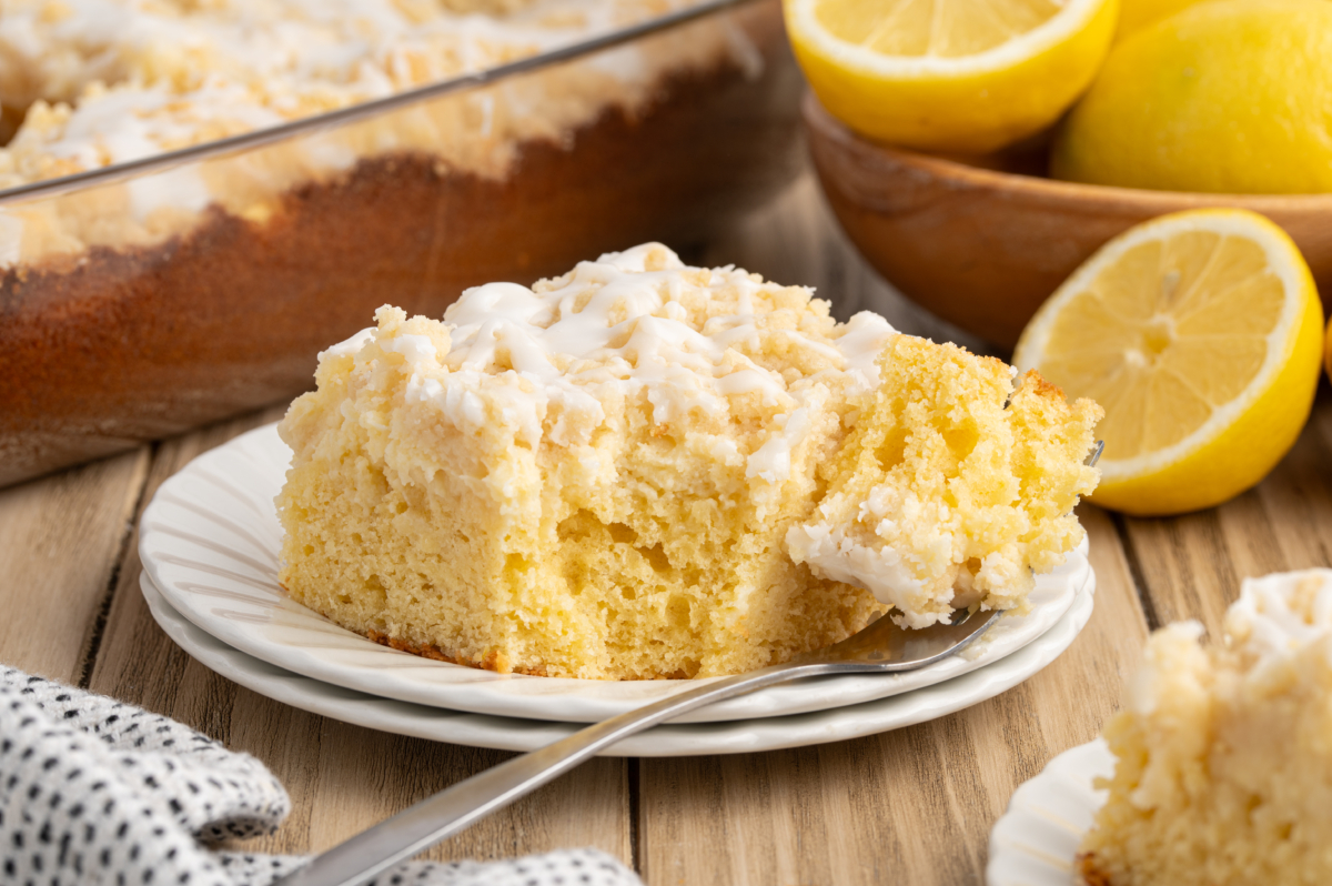 A slice of Lemon Coffee Cake with white icing sits on a white plate, a fork holding a bite. Fresh lemons and a baking dish with more cake appear in the background on a wooden table.
