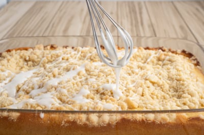 A whisk drizzles white icing over a freshly baked Lemon Coffee Cake crumb cake in a glass baking dish on a wooden table.