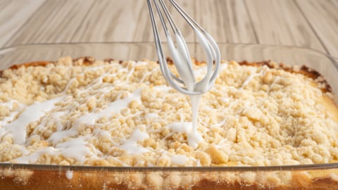 A whisk drizzles white icing over a freshly baked Lemon Coffee Cake crumb cake in a glass baking dish on a wooden table.
