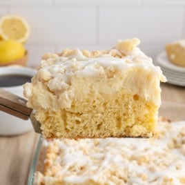 A close-up of a slice of Lemon Coffee Cake with white icing drizzle being lifted from a baking dish, showing its moist, yellow crumb and crumbly topping. Lemons and coffee are in the background.