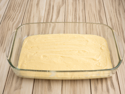 A glass baking dish filled with an even layer of yellow Lemon Coffee Cake batter sits on a light wooden surface, ready to be baked.