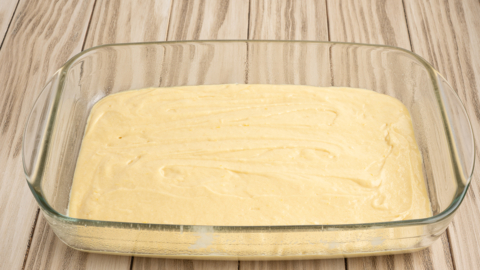 A glass baking dish filled with an even layer of yellow Lemon Coffee Cake batter sits on a light wooden surface, ready to be baked.