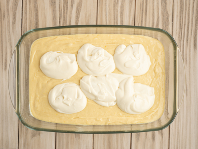 A glass baking dish with a layer of yellow Lemon Coffee Cake batter, topped with six dollops of white creamy mixture, sits on a light wooden surface.